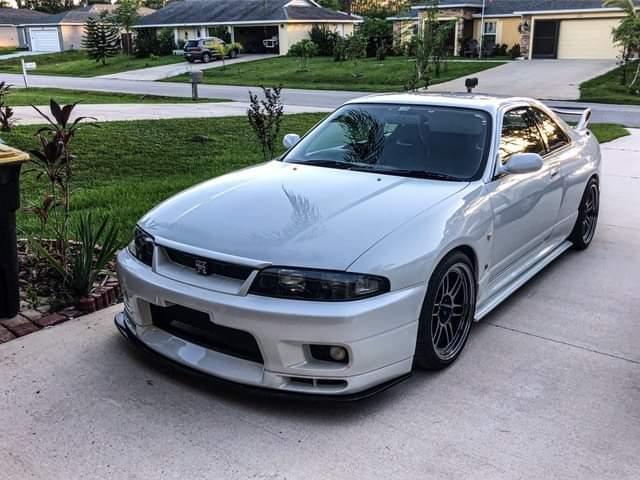 Front side view of a white Nissan Skyline R33 with front lip, side skirts, and aftermarket wheels parked in a driveway