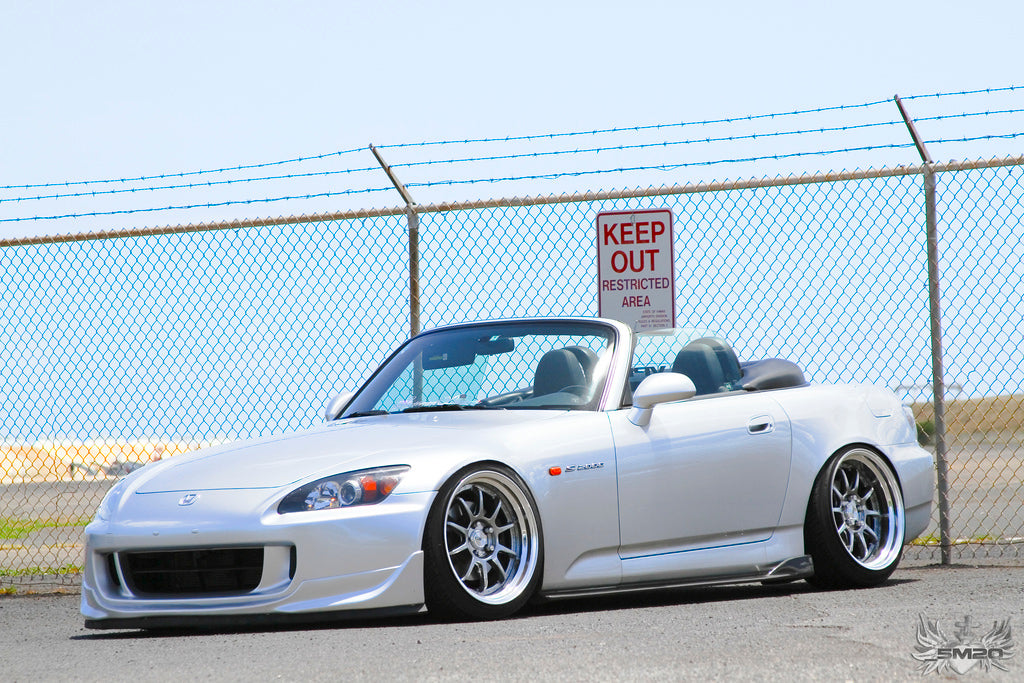 White Honda S2000 convertible with lowered stance, aftermarket wheels, and clean street build styling parked by fence