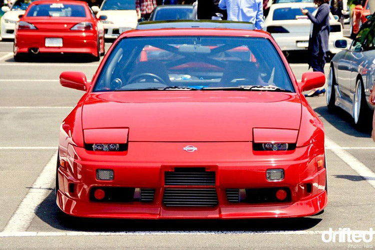 Front view of a red Nissan 180SX with aftermarket front bar, intercooler, and pop-up headlights at a car meet