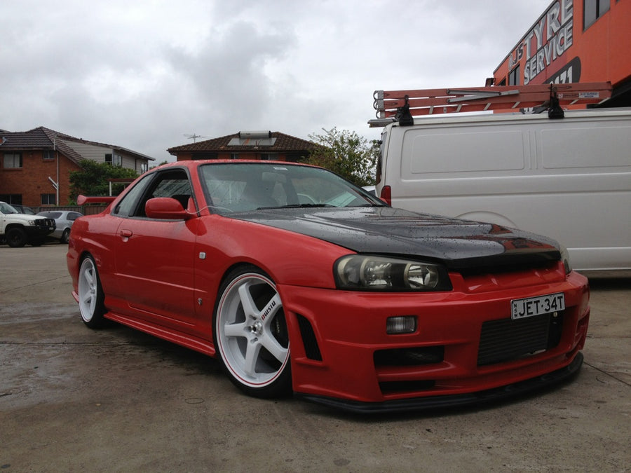 Front angle view of a red Nissan Skyline R34 with JSAI Aero front lip, side skirts, and aftermarket wheels