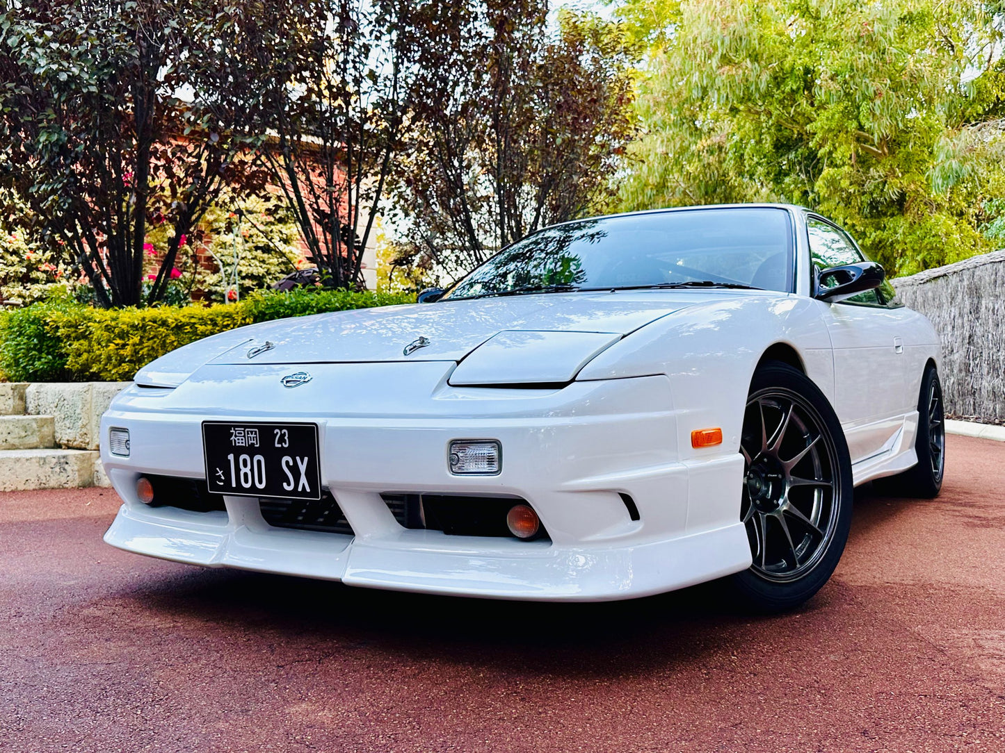 Front angle view of a white Nissan 180SX with front lip, side skirts, and black aftermarket wheels in an outdoor setting
