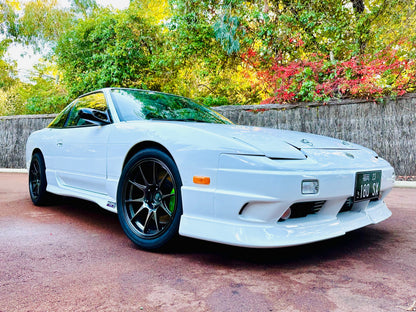 Front side view of a white Nissan 180SX with side skirts, front lip, and black aftermarket wheels parked outdoors
