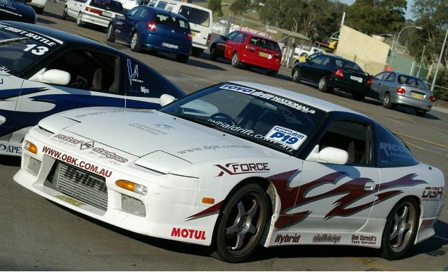 Front side view of a white Nissan 180SX track car with race decals, front-mount intercooler, and aftermarket wheels