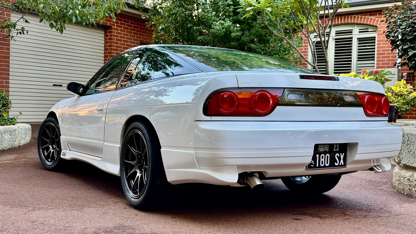 Rear side view of a white Nissan 180SX with side skirts, rear bumper, and black aftermarket wheels parked outdoors