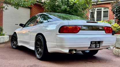 Rear side view of a white Nissan 180SX with side skirts, rear bumper, and black aftermarket wheels parked outdoors