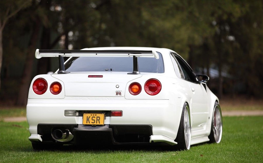 Rear view of a white Nissan Skyline R34 parked on grass with aftermarket rear wing and lowered suspension