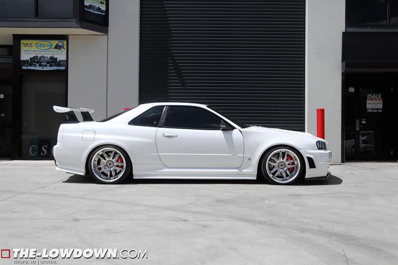 Side profile of a white Nissan Skyline R34 with lowered stance, aftermarket wheels, and rear wing