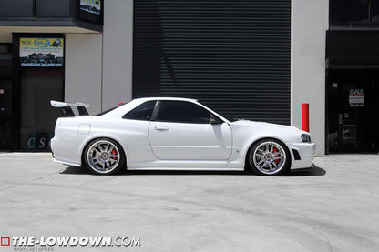 Side profile of a white Nissan Skyline R34 with lowered stance, aftermarket wheels, and rear wing