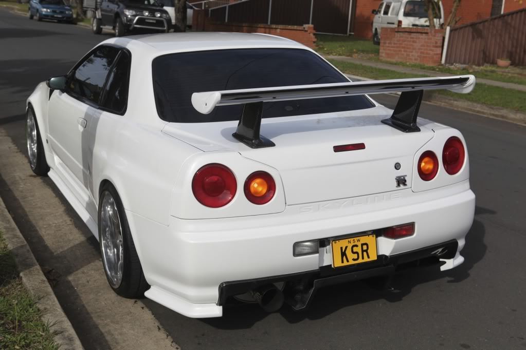 Rear view of a white Nissan Skyline R34 parked on a street with aftermarket rear wing and custom wheels